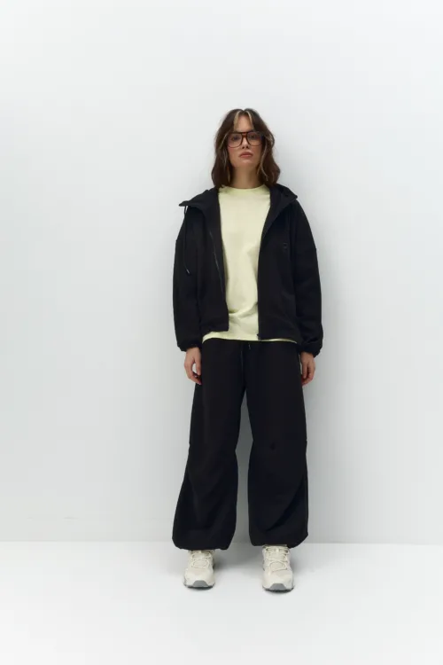 Person standing front-facing against a plain white wall, wearing a black jacket, pale yellow shirt, black pants, white sneakers, and glasses.