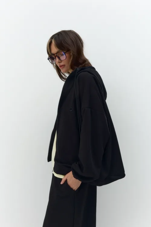 Side view of a person with shoulder-length hair wearing a black hoodie and oversized purple-tinted glasses, looking downward against a white wall.