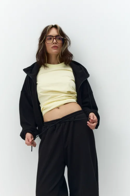 Woman wearing large glasses, a pale yellow top, and a black jacket stands against a white wall, holding a black cord in both hands.