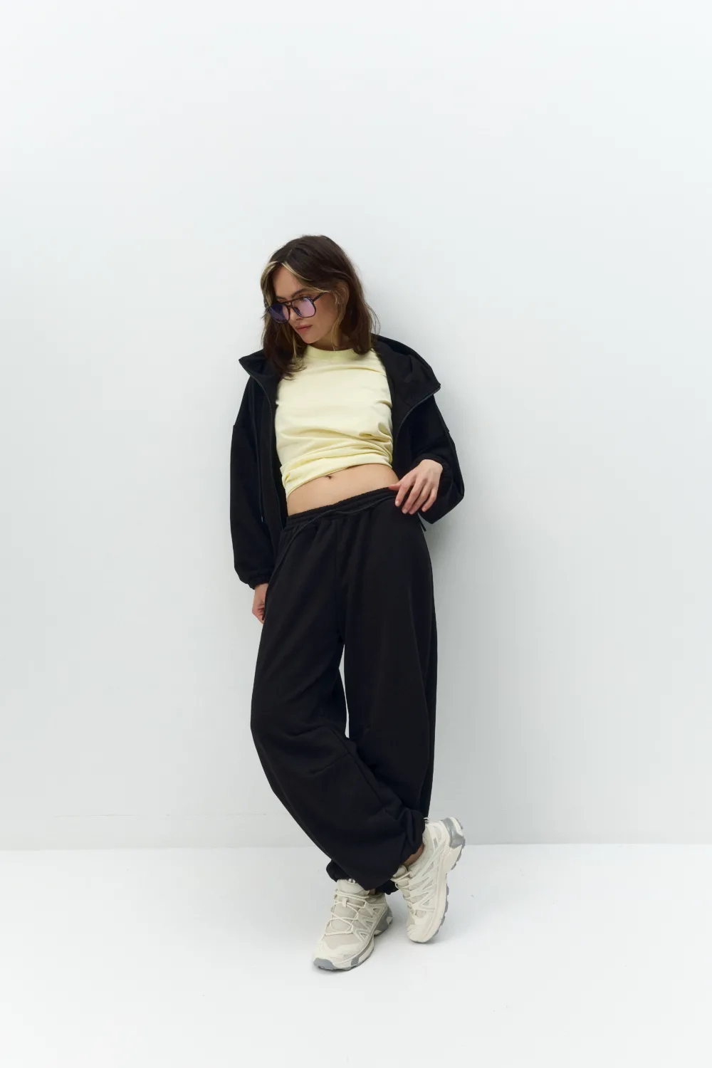 Young woman in a pale yellow top and black tracksuit leans against a white wall, wearing purple sunglasses.