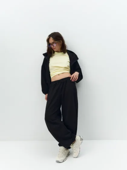 Young woman in a pale yellow top and black tracksuit leans against a white wall, wearing purple sunglasses.