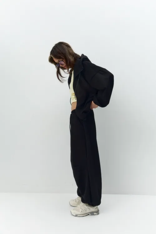 Person wearing a black oversized jacket, cream top, and black pants, bending forward while looking down, with beige sneakers against a plain white background.