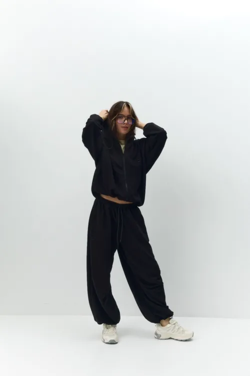 Person in a black hoodie and joggers posing with hands in their hair against a plain white backdrop, wearing light sneakers.