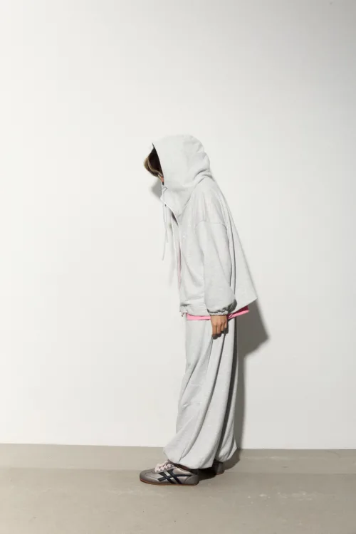 Person in a light gray hoodie and matching sweatpants standing in profile against a plain white wall, pink waistband showing, wearing sneakers.