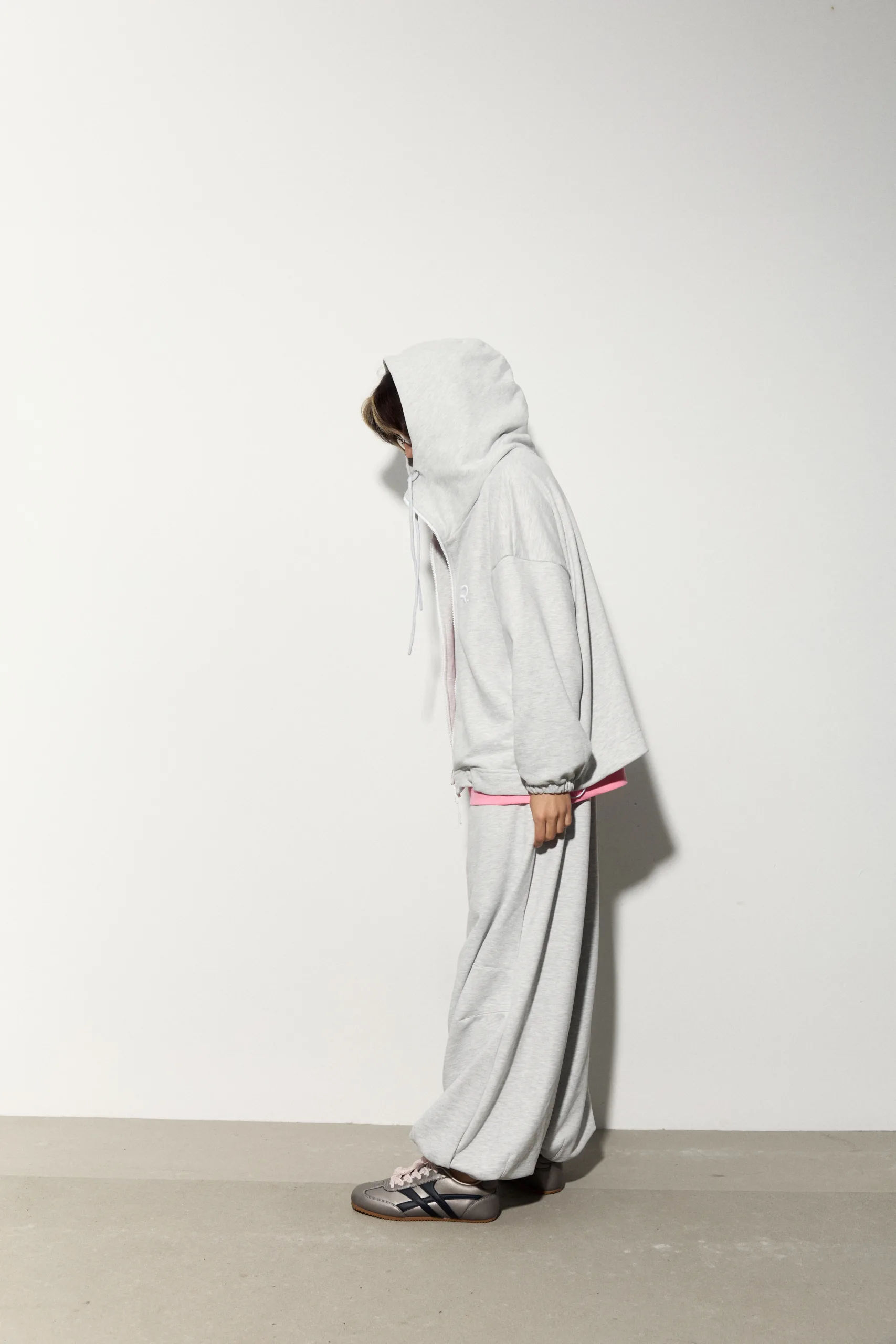 Жіноче oversized худі сірого кольору з футеру діагональ преміум. Вільний силует, блискавка та м’яка фактура для casual образів. - 1C7A1026_Original Худі жіноче oversized сіре на блискавці без утеплення - Person in a light gray hoodie and matching sweatpants standing in profile against a plain white wall, pink waistband showing, wearing sneakers.