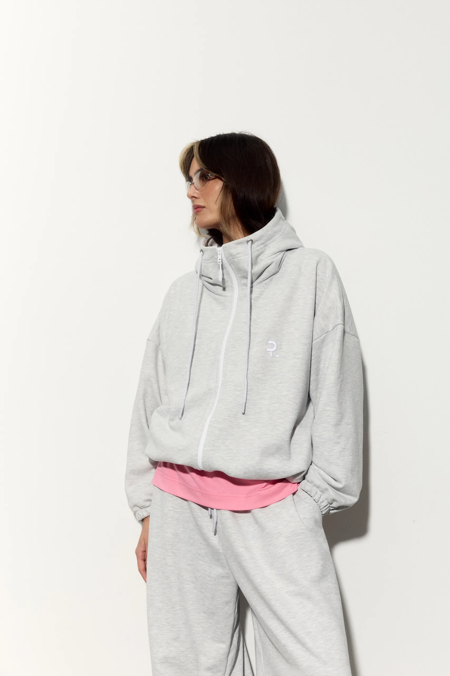 Жіноче oversized худі сірого кольору з футеру діагональ преміум. Вільний силует, блискавка та м’яка фактура для casual образів. - 1C7A1064_Original Худі жіноче oversized сіре на блискавці без утеплення - Person in a light gray hoodie and matching joggers, pink waistband visible, standing in profile against a white wall