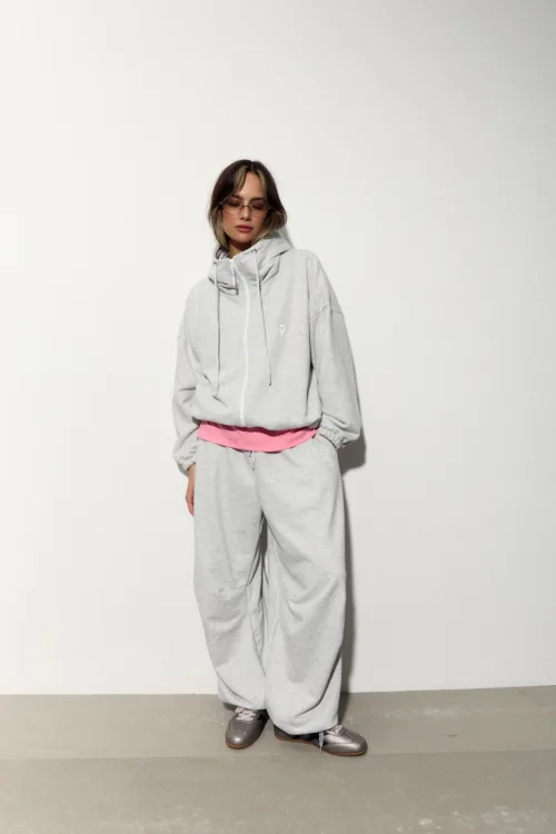 Person in an oversized gray hoodie and matching sweatpants with a pink waistband, standing against a plain white wall.