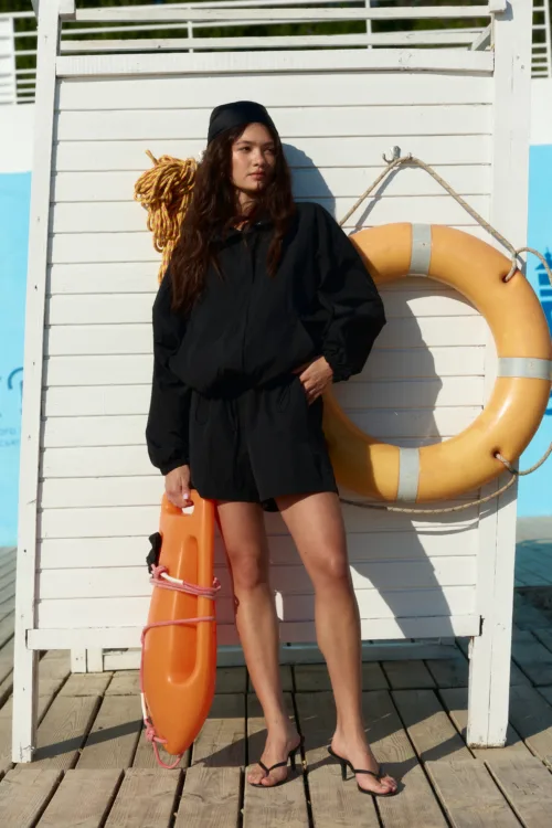 Woman in a black dress stands on a wooden dock beside an orange lifebuoy and coiled rope.