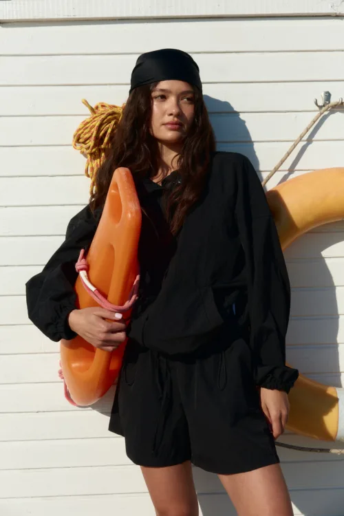 Woman in a black outfit holding an orange life buoy over her shoulder, standing against a white wooden wall with rope in the background.