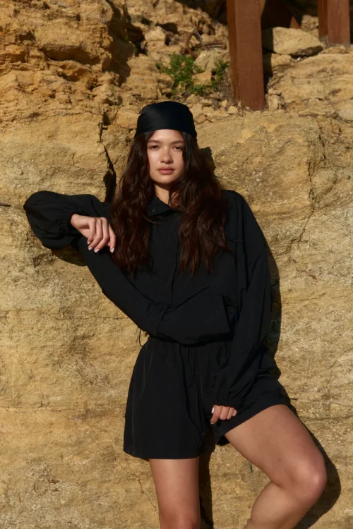 Young woman with long dark hair in a black dress and headscarf posing against a sunlit beige rock wall with her arm resting on the surface.
