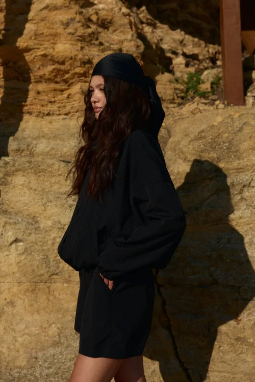Young woman in a black outfit with a headscarf standing against a rugged rocky wall, looking to the left.
