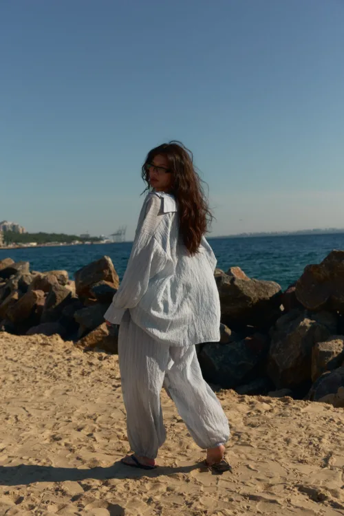 Person walking along a rocky beach, facing away, in a loose white striped outfit with sandals, by the sea under a clear blue sky.
