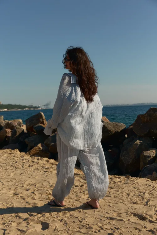 Woman with wavy brown hair in a light striped outfit stands on sandy beach by rocks, gazing toward the water with blue sky above.