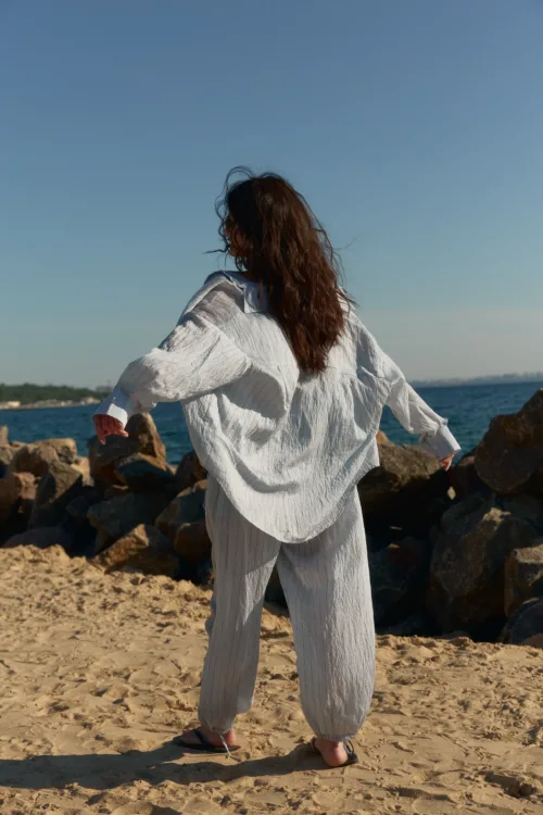 Person seen from behind on a sandy beach, wearing a loose white striped outfit, arms outstretched toward the sea and rocks.