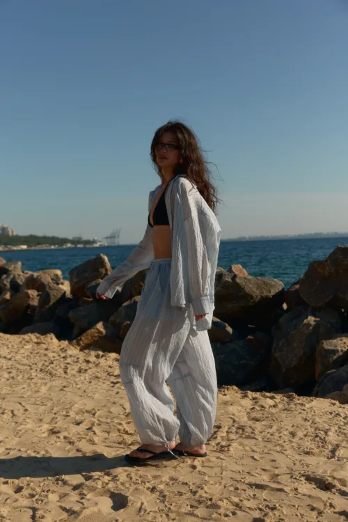 Woman in a striped loose cover-up over a black bikini top stands barefoot on a sandy beach with rocks and the sea behind her.