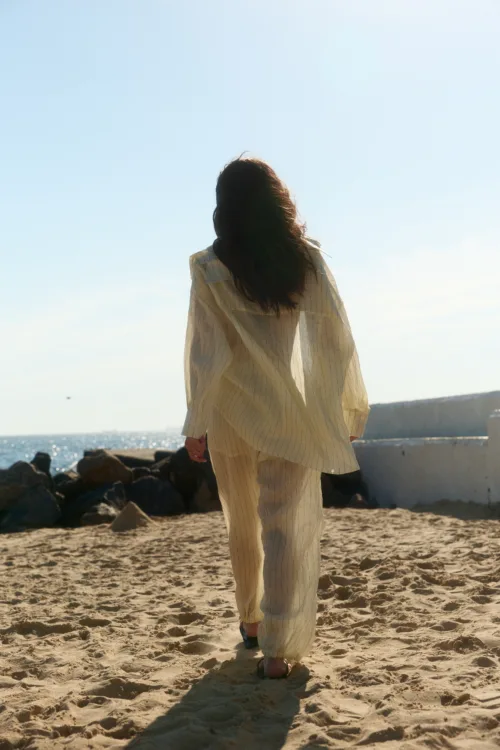 Person seen from behind walking on a sunny beach in a light striped beige outfit, with rocks and the ocean in the background.