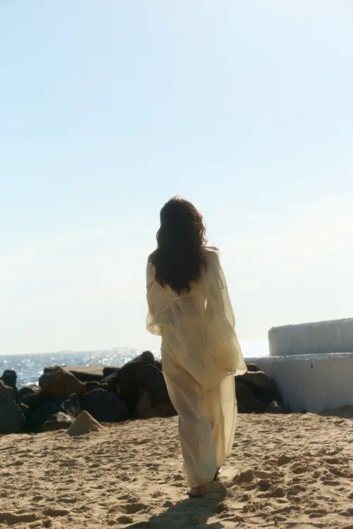 Person seen from behind, walking on a sandy beach in a light-colored dress toward the ocean.