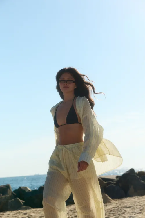 Woman on a sunny beach wearing a black bikini top and striped sheer cover-up, with wind-swept hair and sunglasses, by the rocky shore.