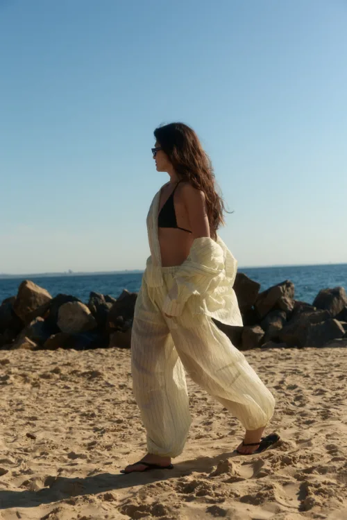 Woman walking along a sandy beach near rocky breakwater, wearing a black bikini top and a light striped cover-up with flip-flops.