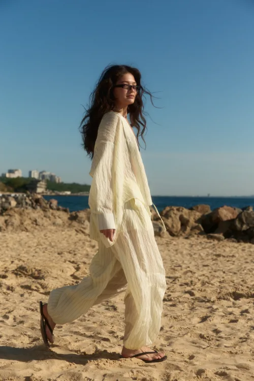 Woman walking along a sandy beach in a cream-colored, striped dress with flip-flops, under a clear blue sky. In the background, rocky shore and distant buildings by the sea.