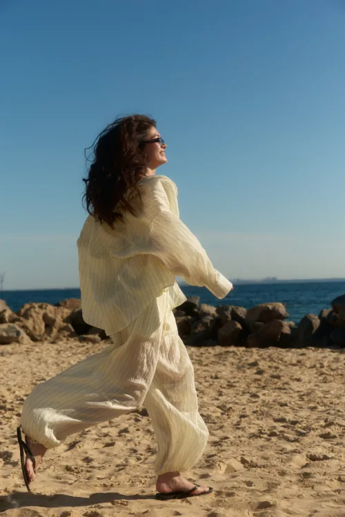 Woman walking along a sandy beach in a flowing cream dress, hair blowing, wearing sunglasses, with the blue sea and sky in the background.