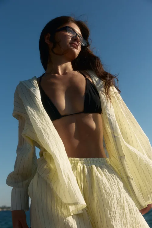 Woman in a black bikini top and pale yellow striped cover-up, standing outdoors against a clear blue sky.