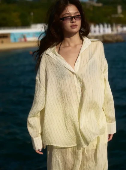 Woman standing by the sea in a cream striped shirt, wearing sunglasses, with wind-blown hair.