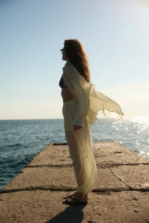 Person standing on a concrete pier by the sea, wearing a light striped outfit and flip-flops, facing left with hair blowing in the breeze.