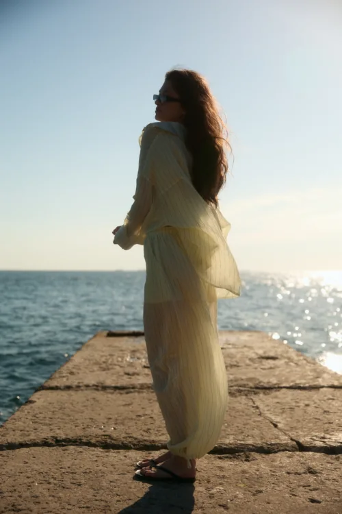 Woman in a pale yellow striped dress and sandals stands on a sunlit pier by the sea, facing left with sunglasses on.