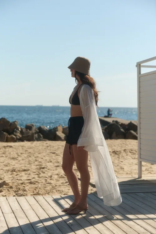 Woman in a black bikini top and white sheer cover-up standing on a wooden boardwalk at the beach with the ocean behind her.