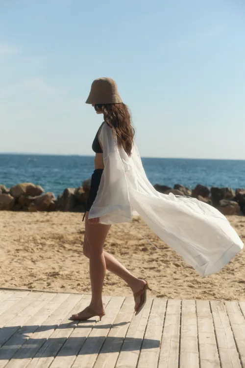 Woman walking barefoot on a wooden boardwalk at the beach, wearing a black bikini, wide-brim hat, and a flowing white cover-up with the sea behind her.