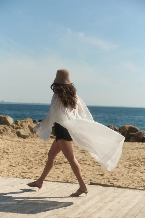 Woman walking on a sandy beach wearing a wide-brim hat and white flowing shawl, with the sea in the background.