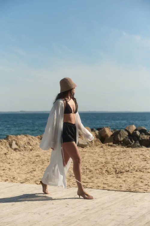 Woman in a black bikini with a white cover-up and tan hat walking along a sandy beach by the sea, near rocks.