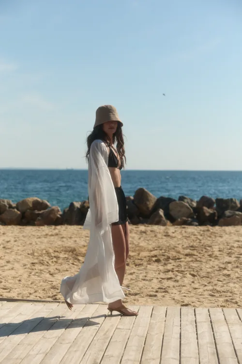 Woman in a black bikini top and sheer white cover-up walks along a wooden boardwalk at the beach, beige sun hat, with the sea and rocks in the background.