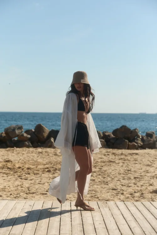 Woman in a black bikini and white sheer cover-up walking along a wooden boardwalk at the beach, wearing a beige hat with the ocean behind her.