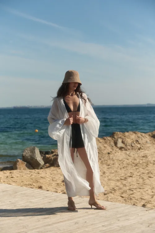 Woman in a black bikini under a sheer white cover-up, wearing a tan bucket hat, standing on a wooden boardwalk by the sea.