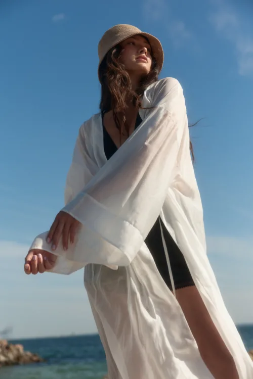 Woman in a light beige sunhat and a loose white cover-up standing by the sea with a clear blue sky behind her; looking off into the distance.