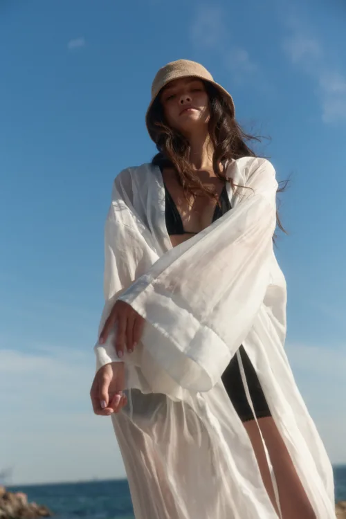 Woman at the beach wearing a straw hat and a white cover-up over a black bikini, against a clear blue sky.