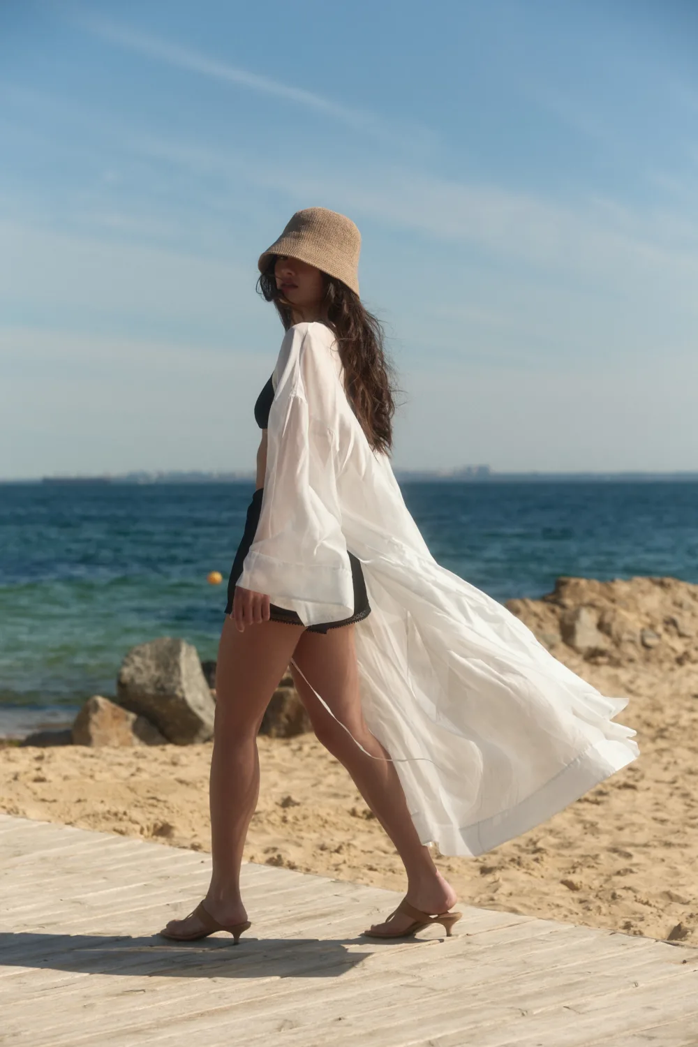 Woman walking along a wooden boardwalk at the beach, wearing a beige hat, white sheer cover-up, and black bikini.