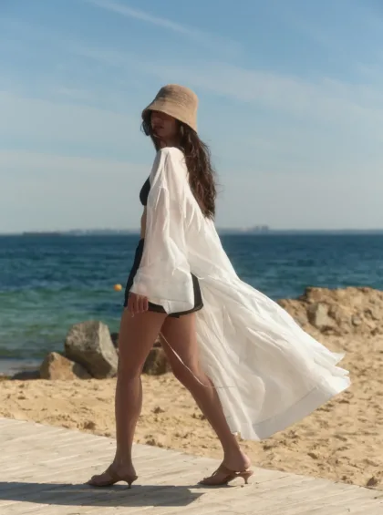 Woman walking along a wooden boardwalk at the beach, wearing a beige hat, white sheer cover-up, and black bikini.