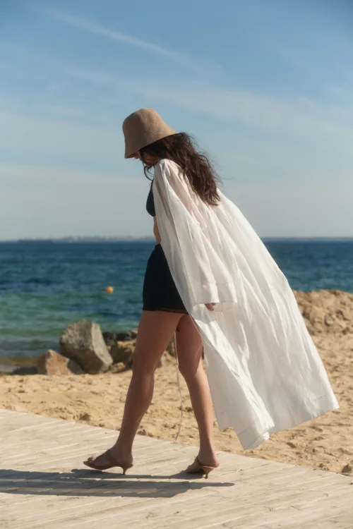Woman walking along a sandy beach in a white flowing cover over a black bikini, with a tan sun hat and bare feet, by the shore.