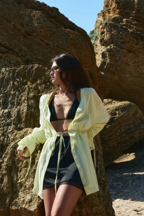 Woman in a black bikini with a pale yellow sheer cover-up, wearing sunglasses, standing against rocky coastal cliffs.