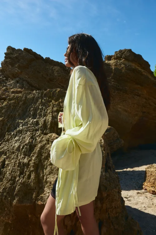Side profile of a person with long dark hair wearing a pale yellow coat, standing beside rugged rocks under a bright blue sky.