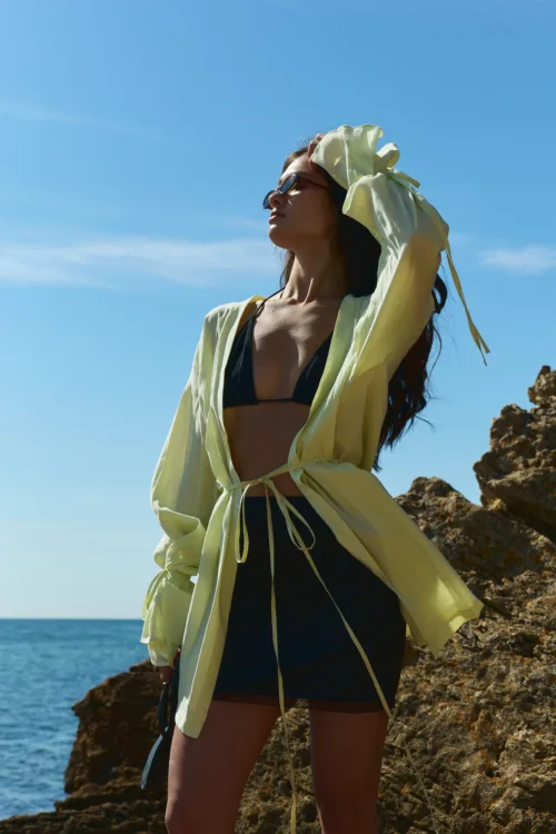 Woman in a pale yellow cover-up over a black bikini, wearing sunglasses, standing on rocky coastline by the blue sea and sky.
