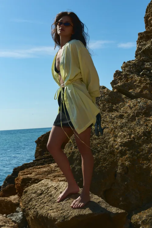 Woman wearing a pale yellow cover-up over a black bikini, barefoot on rocky shore with blue sea and sky behind.