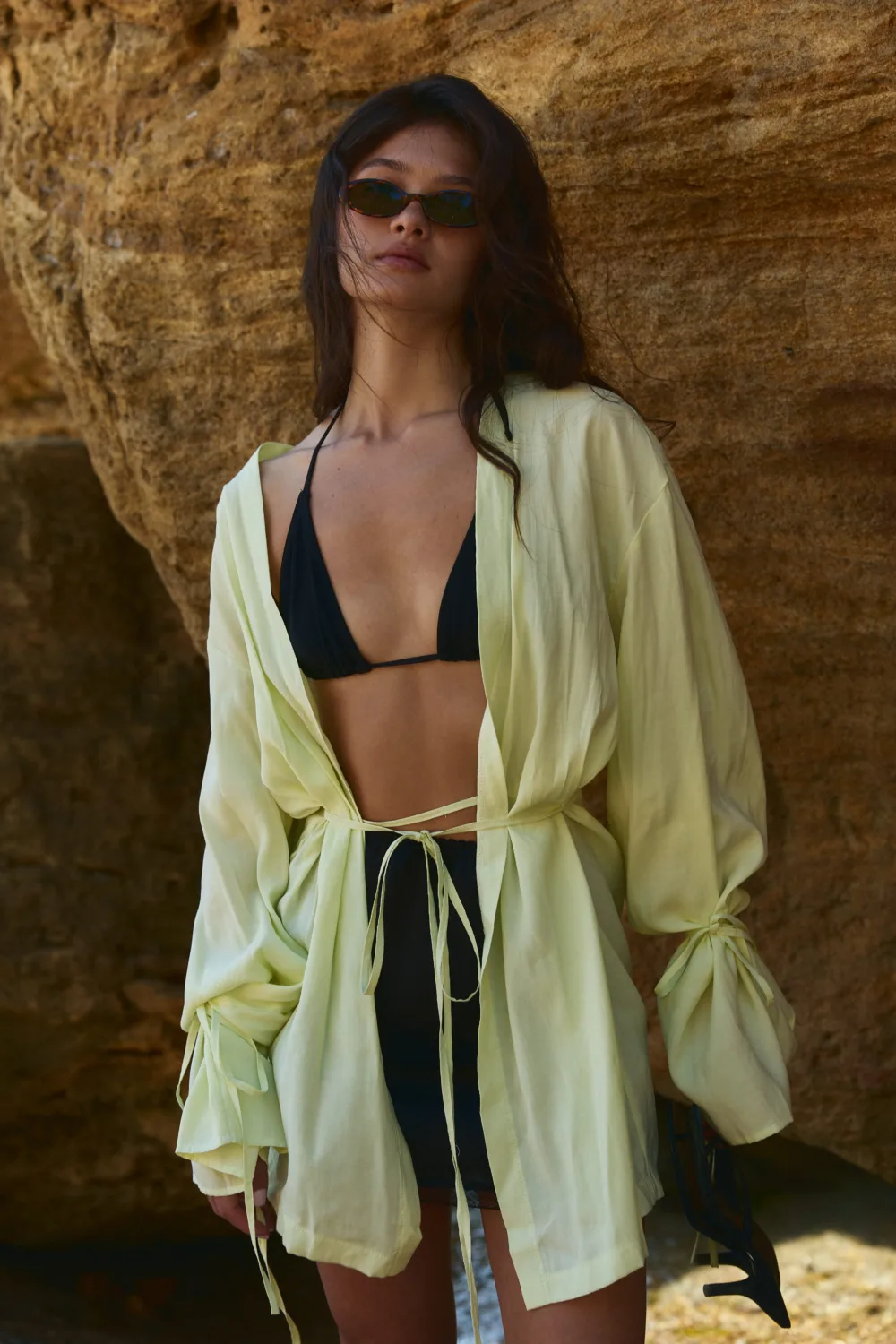 Woman wearing black bikini top under a pale yellow cover-up, sunglasses, standing against a rocky backdrop with wind in her hair.