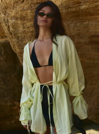 Woman wearing black bikini top under a pale yellow cover-up, sunglasses, standing against a rocky backdrop with wind in her hair.