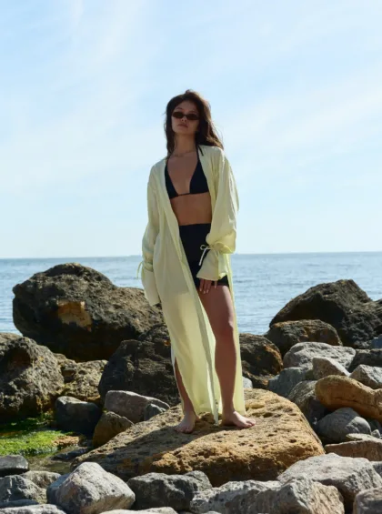 Woman in a black bikini with a light yellow cover-up, barefoot on rocky shore with the ocean in the background, wearing sunglasses.
