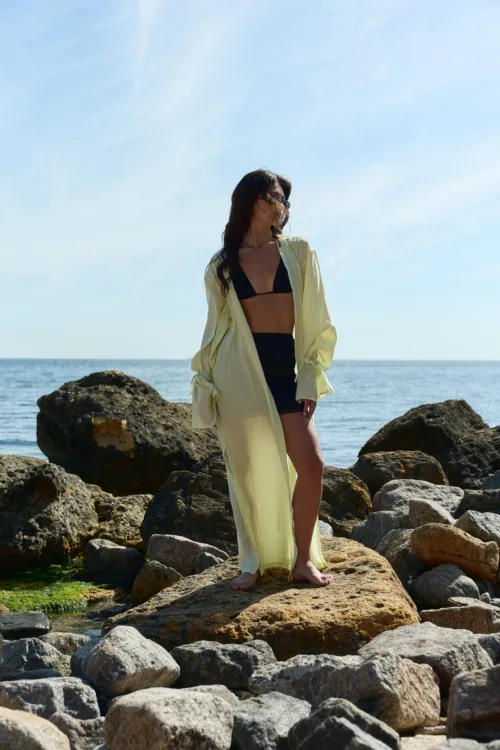 Woman in a black bikini and pale yellow cover-up standing on rocky shoreline with the sea behind her, sunny sky above