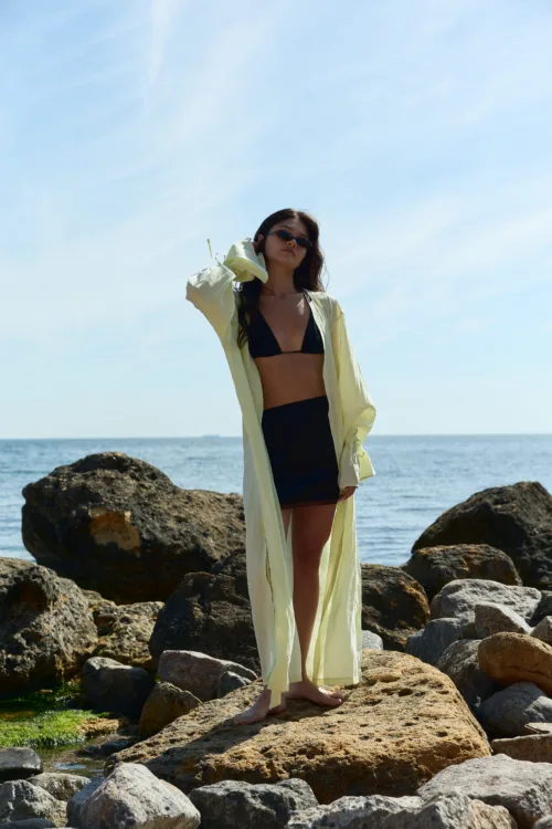 Woman in a black bikini and pale yellow cover-up standing barefoot on rocky shore with the sea in the background and wearing sunglasses against a bright blue sky.